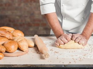 Na imagem vemos uma pessoa, vestindo um dólmã, amassando uma porção de massa, com as duas mãos, sobre uma superfície branca. Ao lado, na mesma superfície em que está fazendo isso, temos um rolo de massa e, ao lado dele, diversões pães, já assados, em formatos diferentes. Ao fundo podemos ver, ainda que desfocado, uma parede de tijolos.