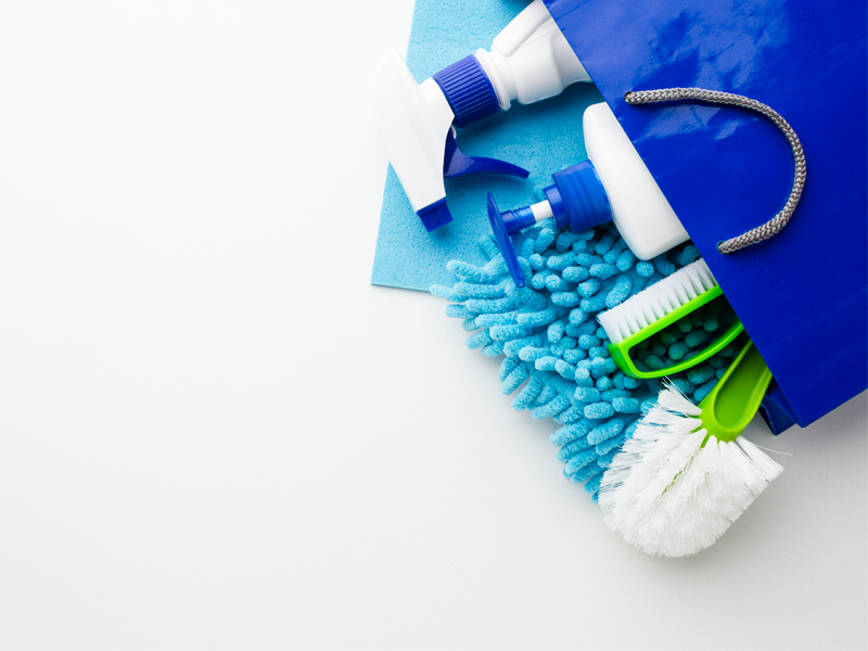 Na imagem vemos uma sacola azul com alça cinza. Dentro dela há diversos itens para limpeza, como uma escova de mão para limpeza, um borrifador, uma escova sanitária e panos. Os itens são azuis e brancos, com exceção da escova de mão para limpeza e da escova sanitária, que possuem cerdas brancas e detalhes em verde. O fundo da foto é claro.
