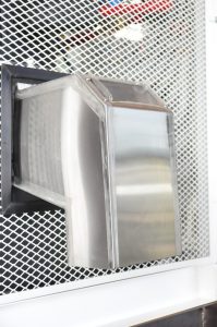 Na imagem é possível ver a saída de gelo da máquina. Ela é retangular e prateada, atrelada à um quadrado escuro com telas brancas ao redor.