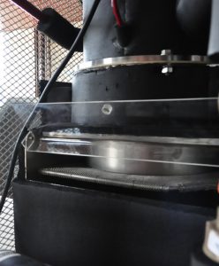 Imagem interna da máquina, mostrando o tanque da mesma, em cor predominantemente preta e em close. É possível ver dois cabos, um preto e outro vermelho, e grades da máquina ao fundo.