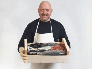 A imagem mostra um homem caucasiano vestindo jaleco e segurando uma caixa com diversos frutos do mar, como peixes pequenos e crustáceos. Ele está em um ambiente vazio com parede cinza.