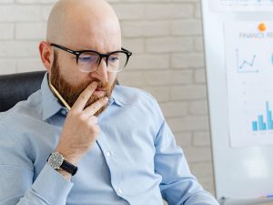 A imagem mostra um homem branco, calvo, com barba ruiva, sentado em uma cadeira. Ele usa óculos de grau, veste uma camisa azul e está com uma expressão pensativa. Ao fundo está um quadro com gráficos azuis.