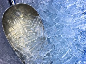A imagem mostra um punhado de gelo em tubo sobre uma superfície plana. Há uma concha prateada com uma pequena quantidade do produto sobre ela.