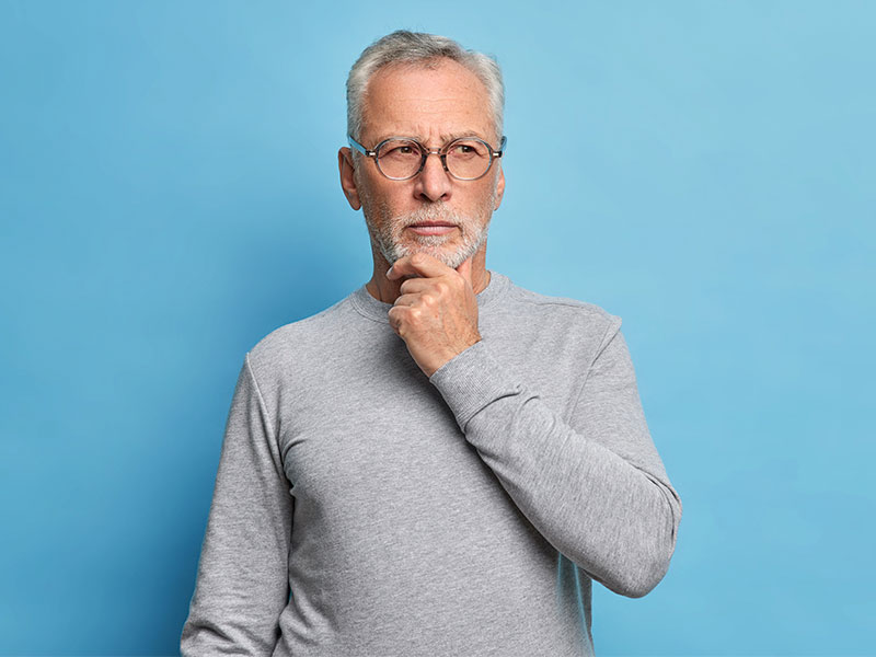 A imagem mostra um homem idoso, com pele branca e cabelos grisalhos. Ele veste um suéter cinza e está com uma expressão pensativa no rosto. O fundo da imagem é azul.