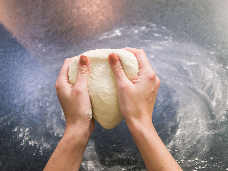 A imagem mostra duas mãos com pele branca e unhas pintadas de salmão segurando uma massa de pão. Ao fundo está uma superfície de granito preto com farinha sobre ela.