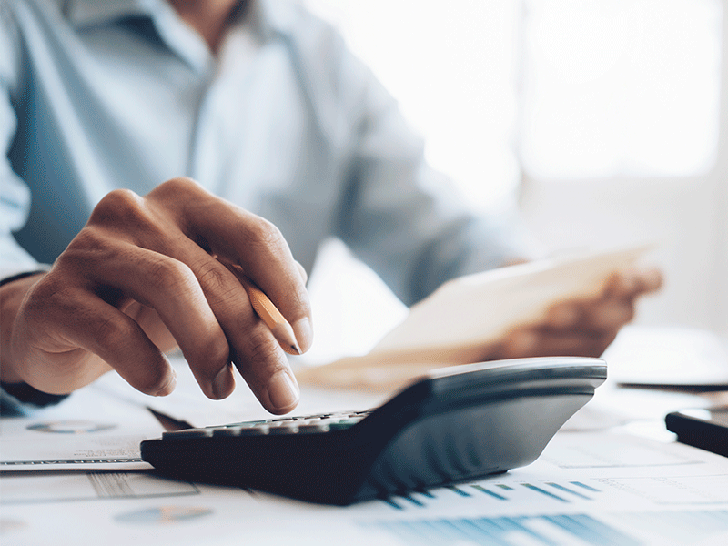 A imagem mostra um close em uma mão que segura um lápis e digita em uma calculadora. Ao fundo, desfocado, está um homem, que veste camisa azul e segura alguns papéis.