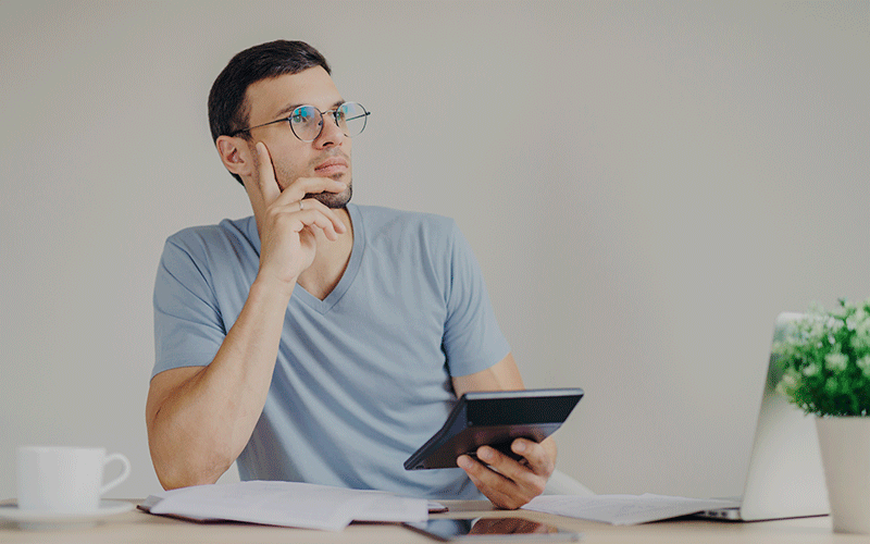 A imagem mostra um homem sentado atrás de uma mesa. Ele está apoiado em uma das mãos com o rosto pensativo. Sobre a mesa estão vários papéis e um notebook. O homem segura uma calculadora. O fundo da imagem é uma parede branca.