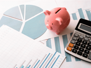 A imagem mostra um cofrinho de porco, uma calculadora e diversos papéis com gráficos sobre uma mesa, fazendo alusão ao financiamento de uma máquina de gelo industrial.