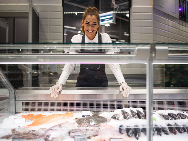 A imagem mostra uma mulher atrás de um freezer expositor com diversos peixes.