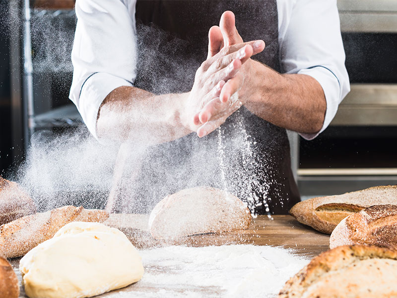 A imagem mostra um homem de avental mexendo com massas de pão sobre uma mesa que já contém pães assados.